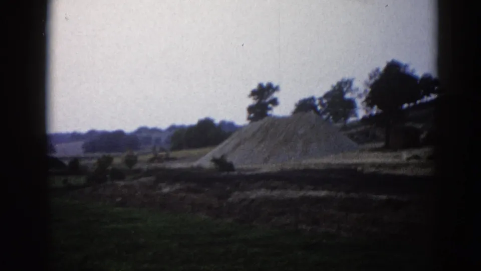 Large conical sand or gravel pile at a rural Illinois construction site, circa 1962, on 8mm film with corner vignetting and a pale overcast sky.