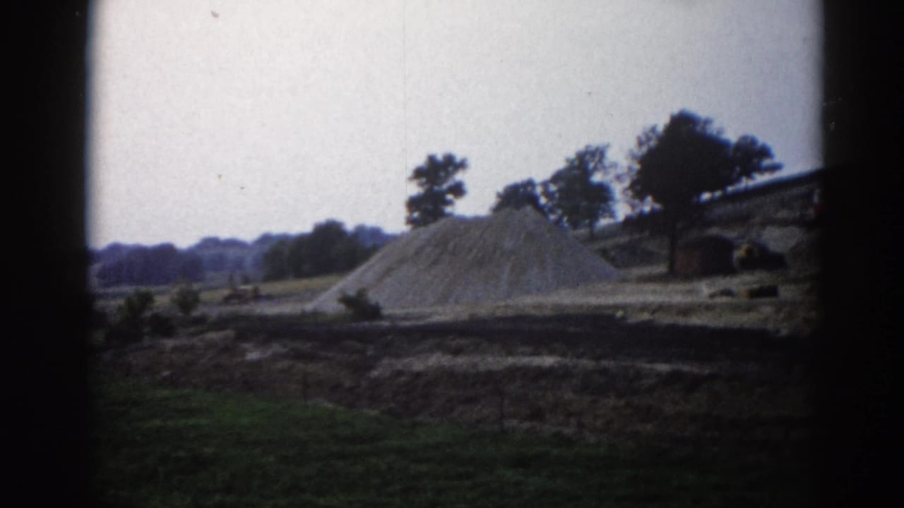 Wide view of a sand pile on a construction site with a tree-lined horizon, 8mm film with corner vignetting.