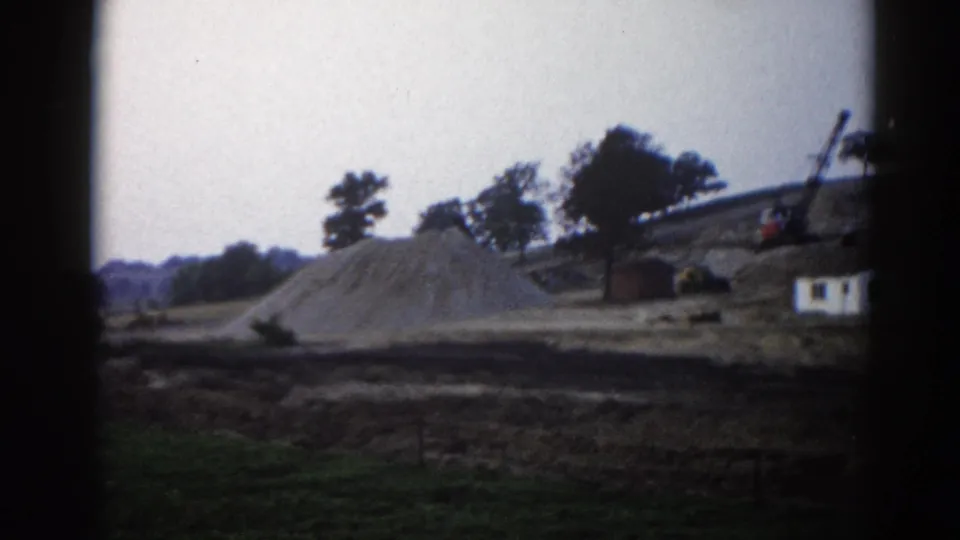 Rural Illinois construction site, circa 1962: sand pile, white site office, crane arm, and heavy equipment against a tree-lined horizon, on 8mm film.