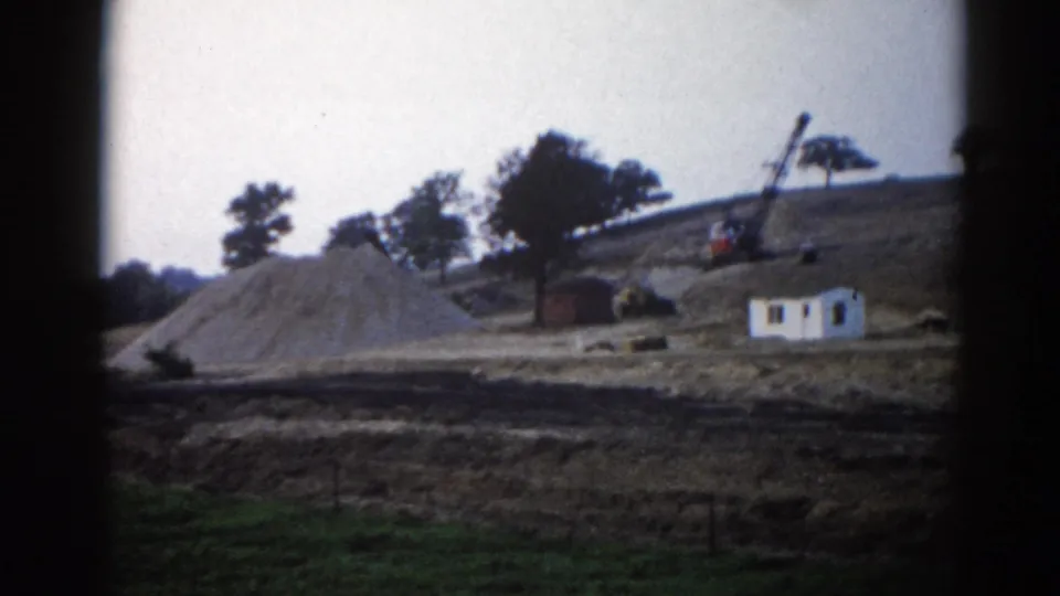 Rural Illinois construction site, circa 1962: sand pile, white site office, crane arm, and construction machinery against a tree line, on 8mm film.