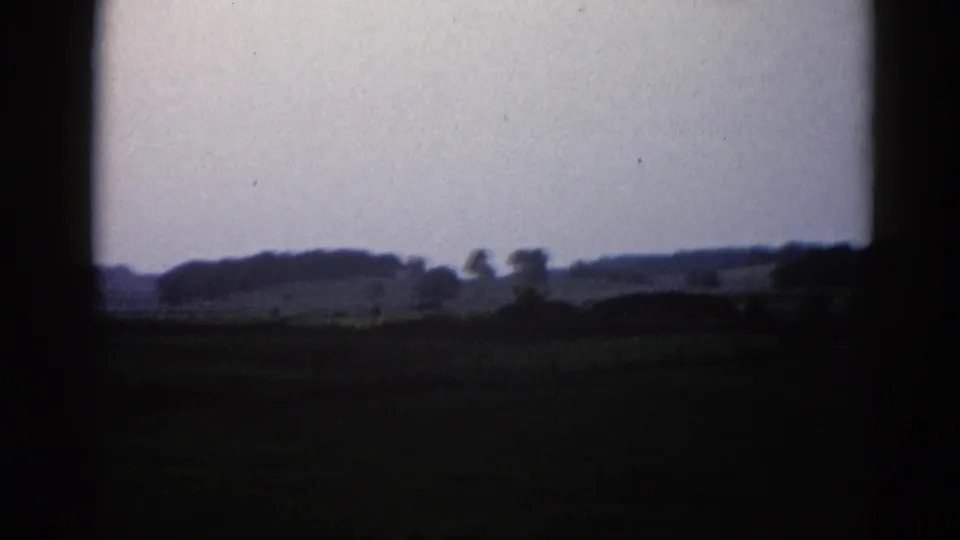 Rural Illinois landscape circa 1962, flat fields and a distant tree line under a heavy overcast sky, captured on 8mm home-movie film.