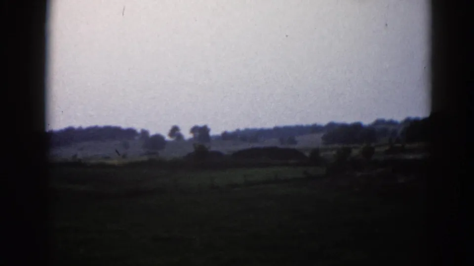 Wide view of rural Illinois fields circa 1962, under a pale overcast sky with deciduous trees at the horizon, captured on 8mm home-movie film.