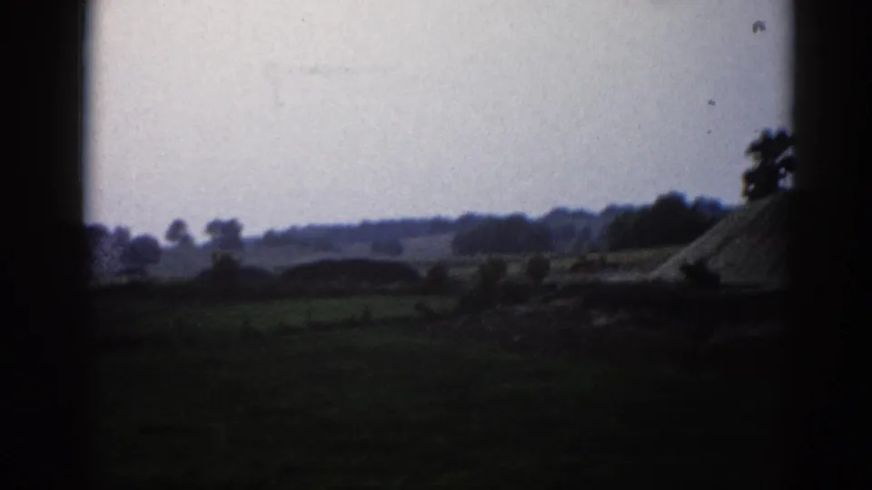 Rural Illinois landscape circa 1962 with a prominent gravel or aggregate mound mid-ground, flanked by trees, under a heavy overcast sky on 8mm film.