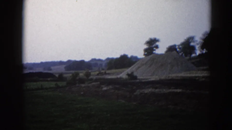 Closer view of a large aggregate mound in rural Illinois circa 1962, trees to the right, pale overcast sky, shot on 8mm home-movie film.