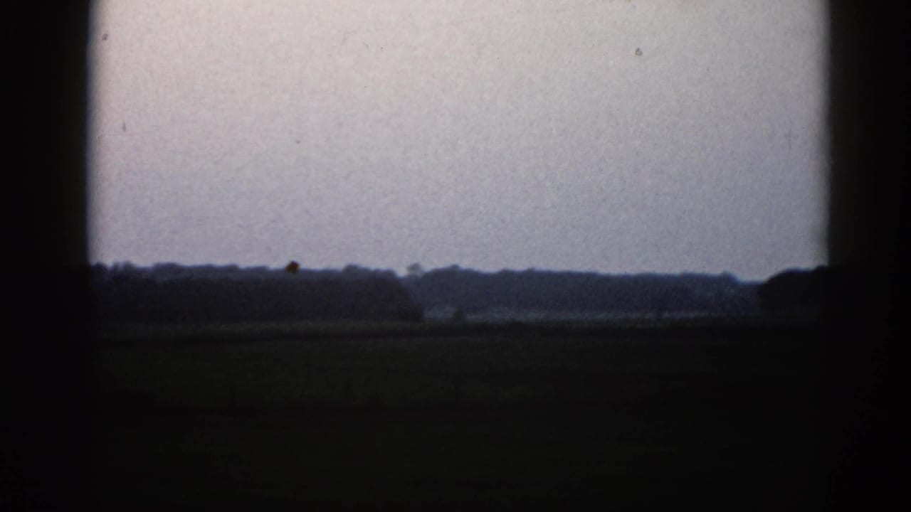 Silhouetted treeline and farm structure against a pale overcast sky, with a dark foreground field and heavy 8mm film borders.