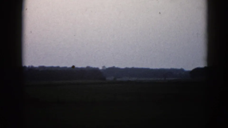 8mm home movie frame from 1962 Illinois showing a silhouetted treeline and barn against a gray overcast sky, flat dark field below.