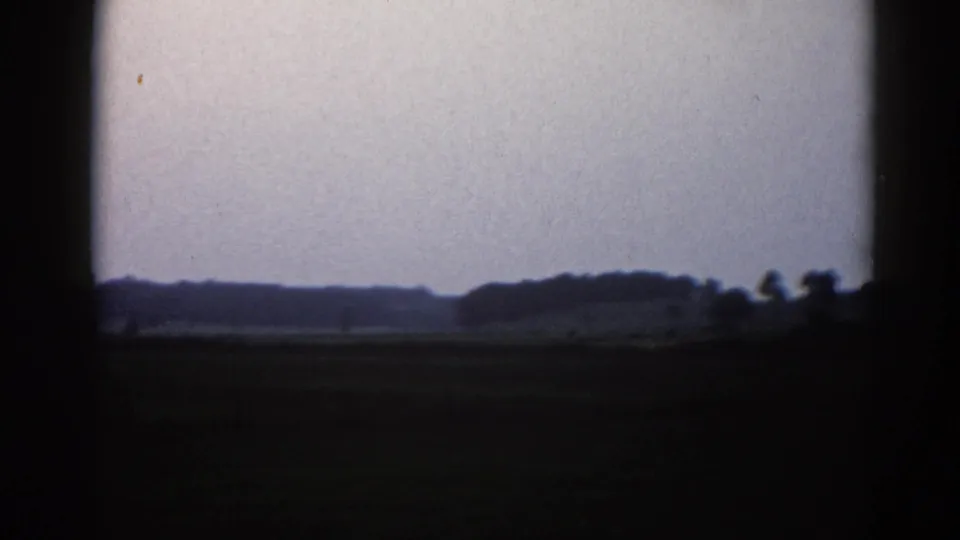 8mm footage from 1962 Illinois showing a low horizon with silhouetted farm buildings and treeline under a pale lavender-gray sky.