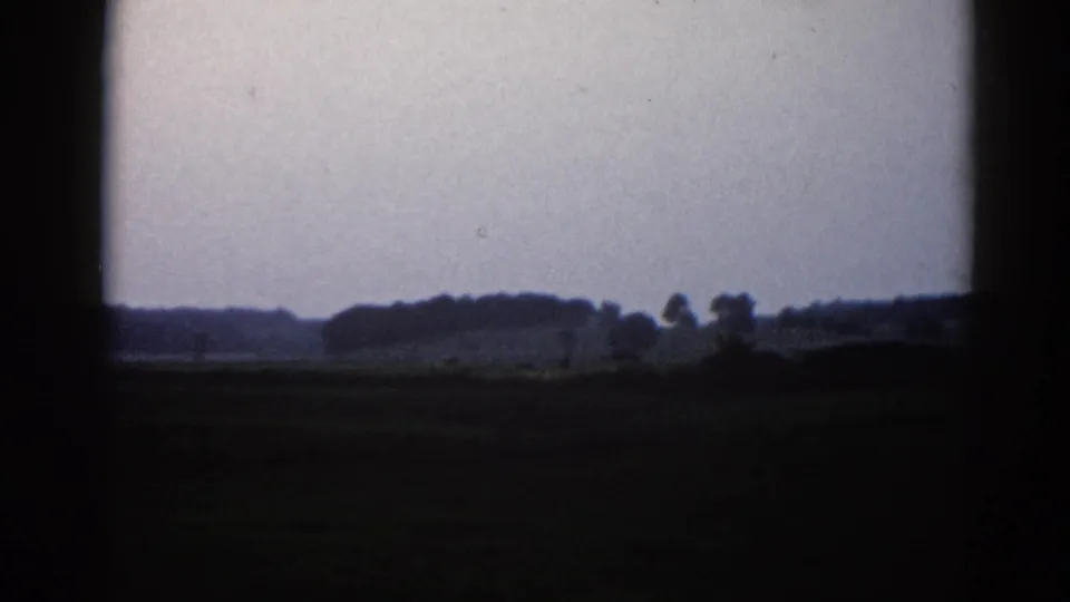 Final frame of 1962 Illinois 8mm pan showing treeline, possible barn or silo silhouettes, and a muted lavender-gray overcast sky.