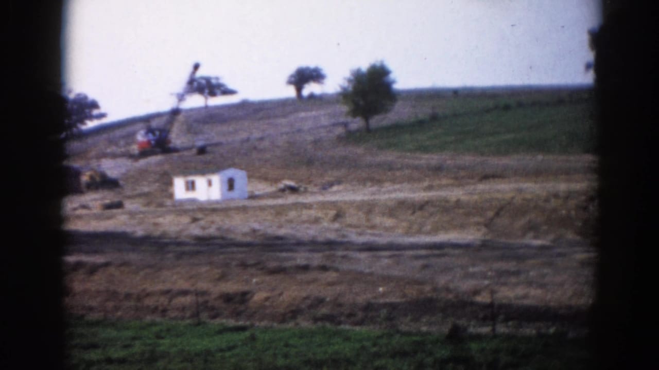 Graded rural hillside with white site trailer and construction equipment, shot on 8mm film with gate vignetting.