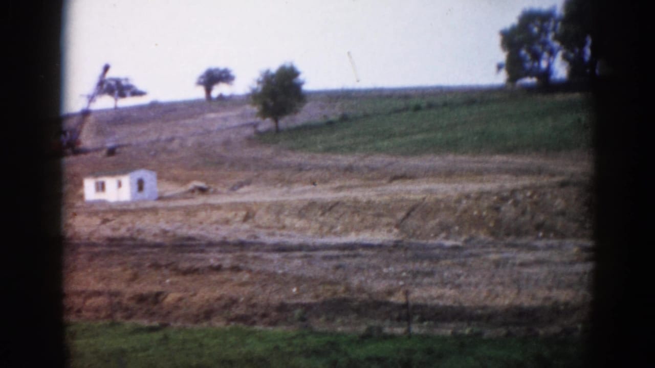 Same rural hillside scene: white site trailer and construction equipment on graded slope, 8mm vignette framing.