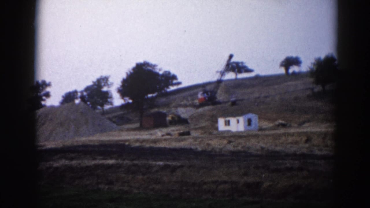 1962 Illinois construction site: small white building and crane on a graded hillside with large earthwork mounds, captured on 8mm home movie film.