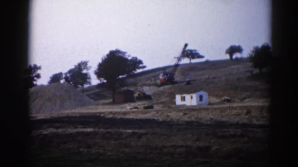 1962 Illinois construction site: small white building and crane on a graded hillside with large earthwork mounds, captured on 8mm home movie film.