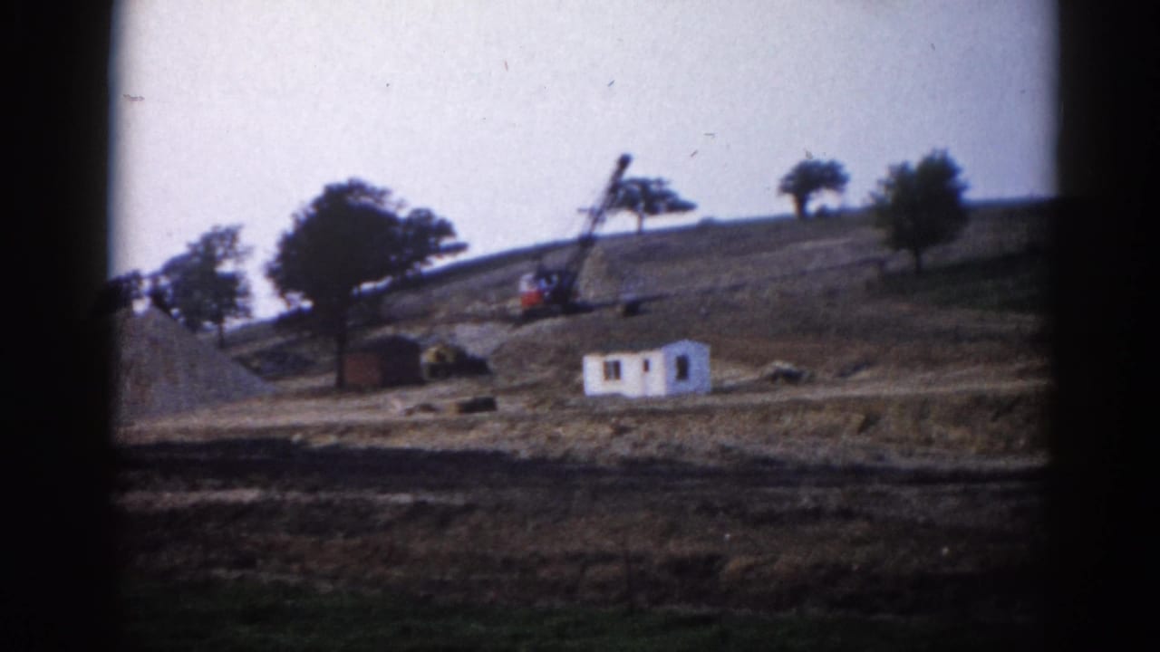 1962 Illinois construction viewed from a distance: crane operating on a dirt hillside beside a white structure, trees silhouetted against a pale sky.