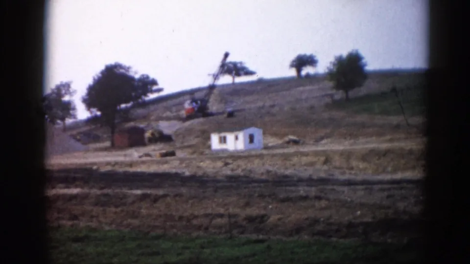 1962 Illinois construction site: orange-bodied crane beside a white building on a graded hillside, green grass visible at lower edge of frame.