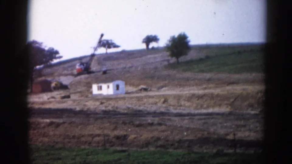 Closing frame of 1962 Illinois construction scene: crane and white building on earthworked hillside, green foreground grass, trees lining the ridge.