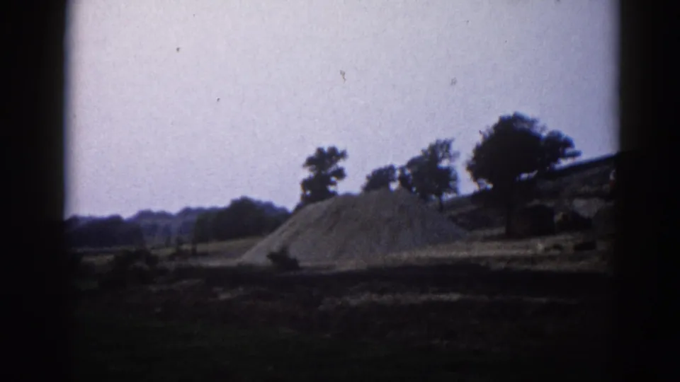 8mm home movie frame circa 1962, Illinois — large conical gravel aggregate mound on a rural construction site with deciduous trees on the ridge.