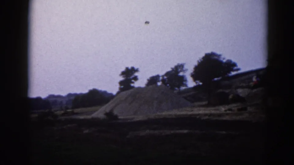 8mm film frame circa 1962, Illinois — gravel pile and sparse treeline under pale overcast sky at a rural construction site.