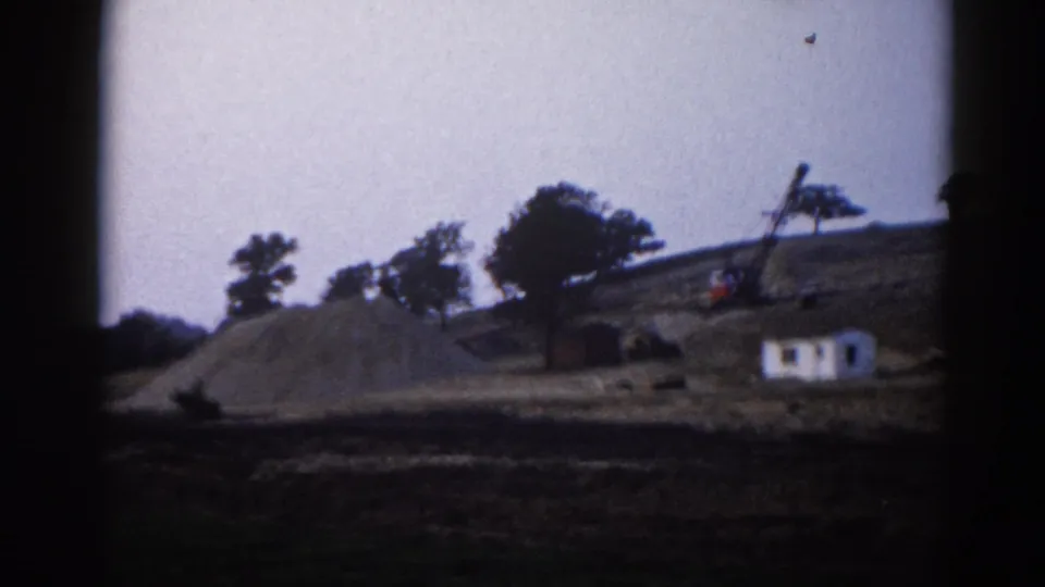 8mm home movie circa 1962, Illinois — construction site showing gravel pile, white site trailer, and crane equipment on a disturbed hillside.