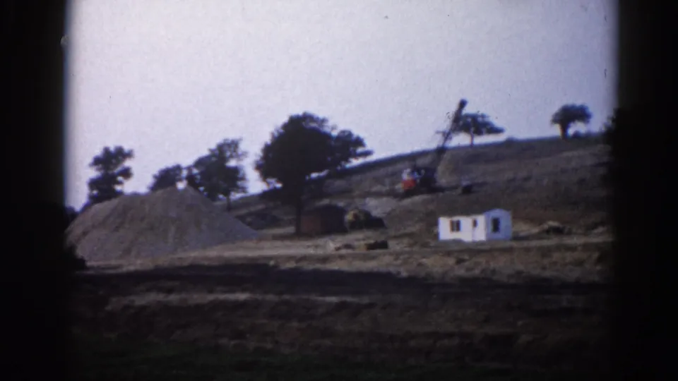 8mm film circa 1962, Illinois — wider view of rural construction site showing white trailer, crane arm, large gravel pile, and treeline.