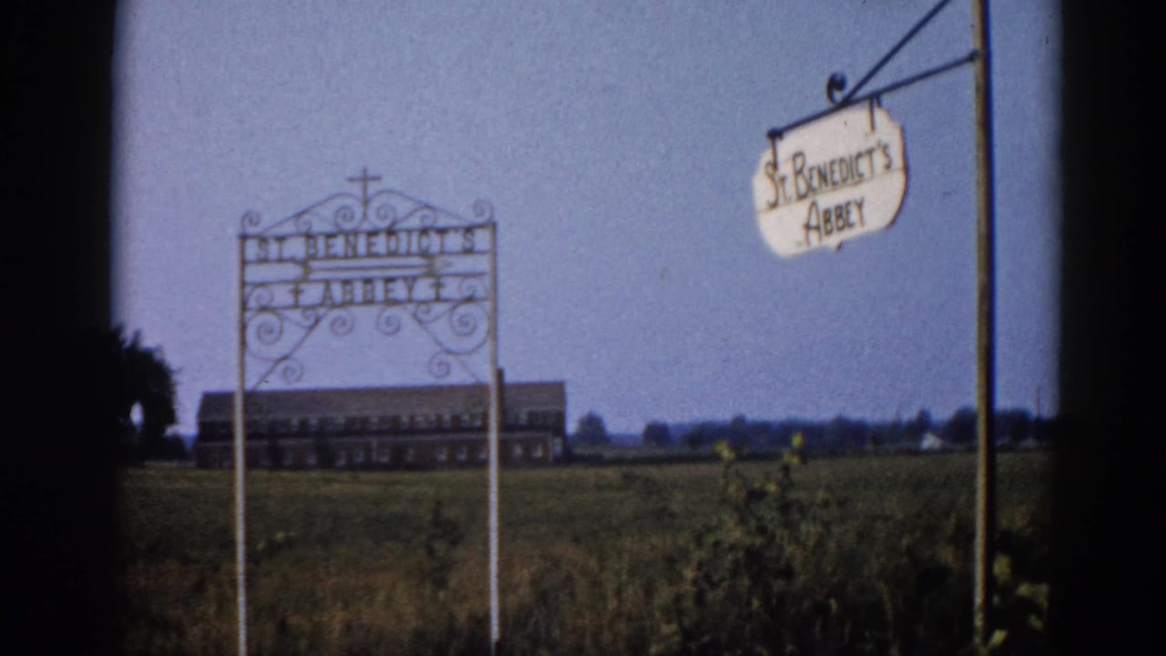 Wrought-iron entry signs reading St. Benedict's Abbey in rural Illinois, circa 1962, with low roadside vegetation, flat agricultural fields, and a large building in the far distance.
