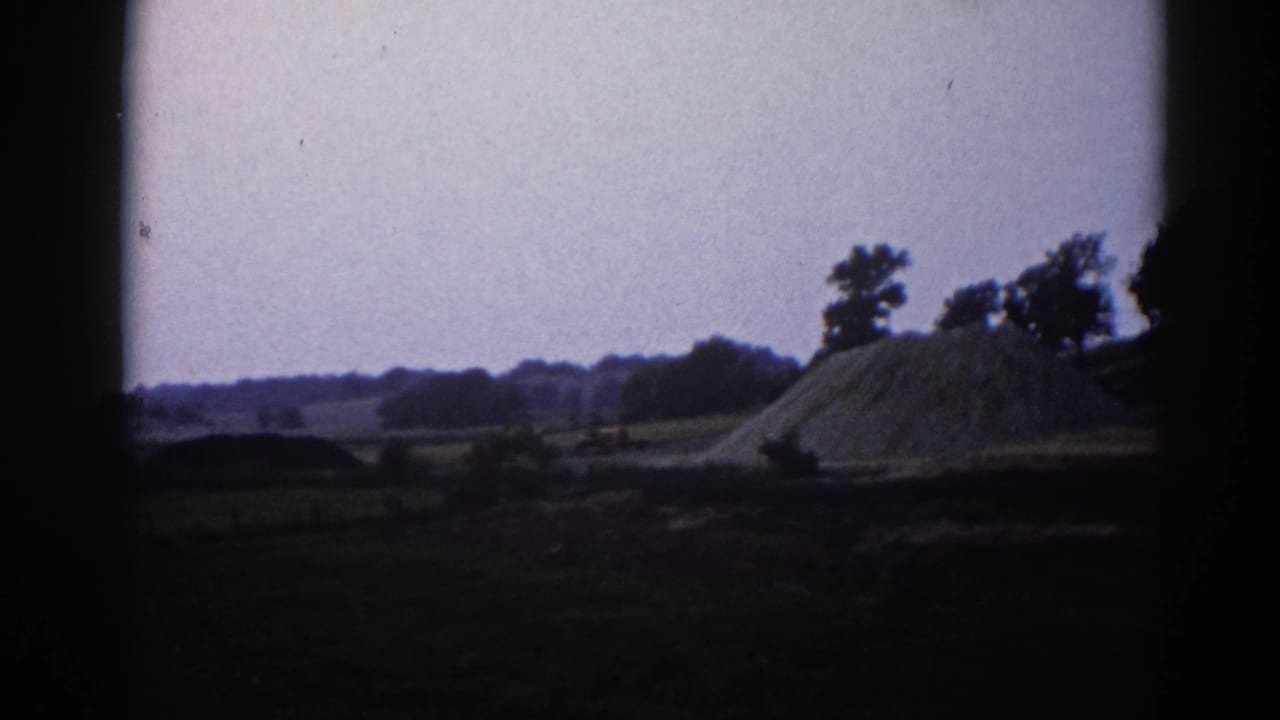 Wide shot of a large conical dirt or gravel pile in a flat rural Illinois landscape, with trees on the horizon and a pale hazy sky.