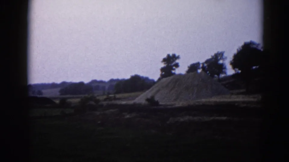 Large dirt or gravel pile in rural Illinois, circa 1962; pale blue-lavender sky and a line of deciduous trees frame the mound under visible 8mm film grain.