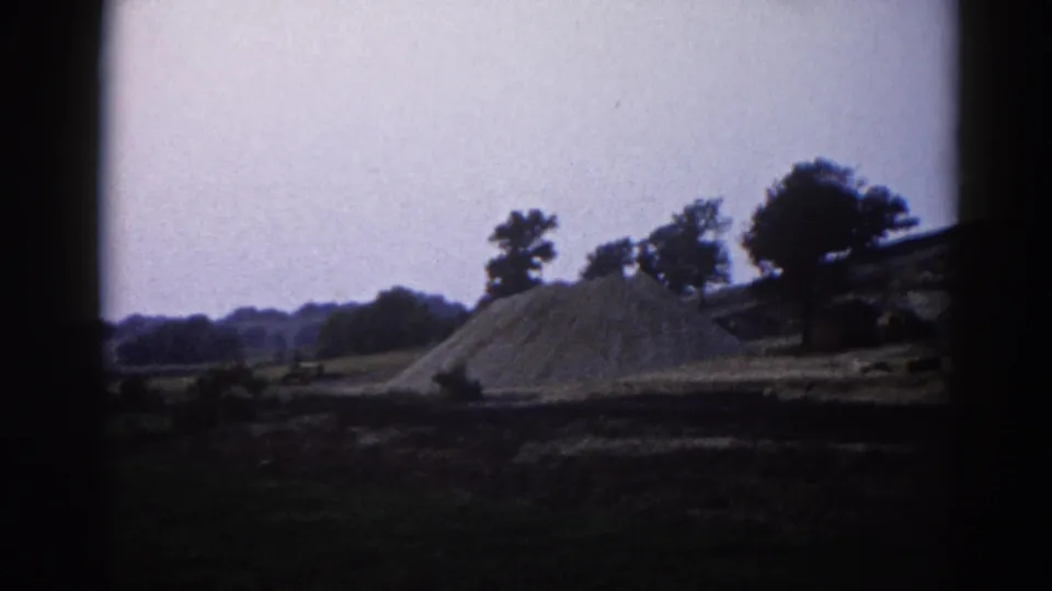 A large earthen mound in a flat Illinois rural field, circa 1962, with trees silhouetted on the right and a hazy overcast sky; slight 8mm vignetting at frame edges.