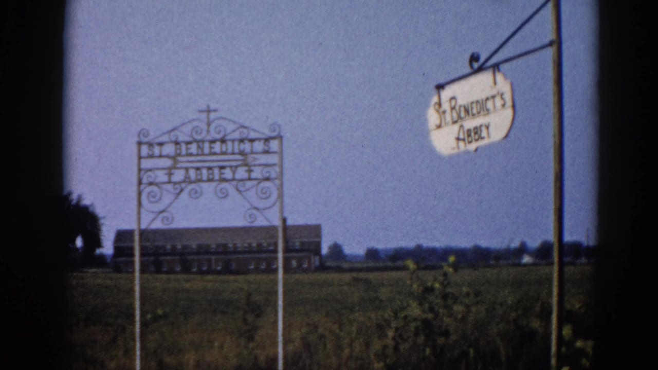 Wrought-iron entrance sign reading St. Benedict's Abbey with scrollwork and cross finial, hanging pendant sign, and brick institutional building in distance; Illinois, 1962, 8mm.