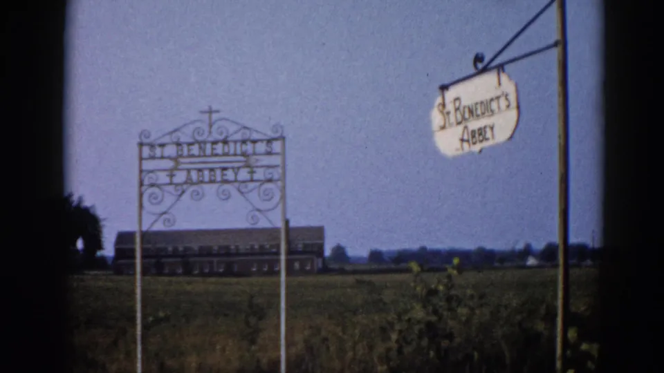 Wrought-iron entrance sign reading St. Benedict's Abbey with scrollwork and cross finial, hanging pendant sign, and brick institutional building in distance; Illinois, 1962, 8mm.