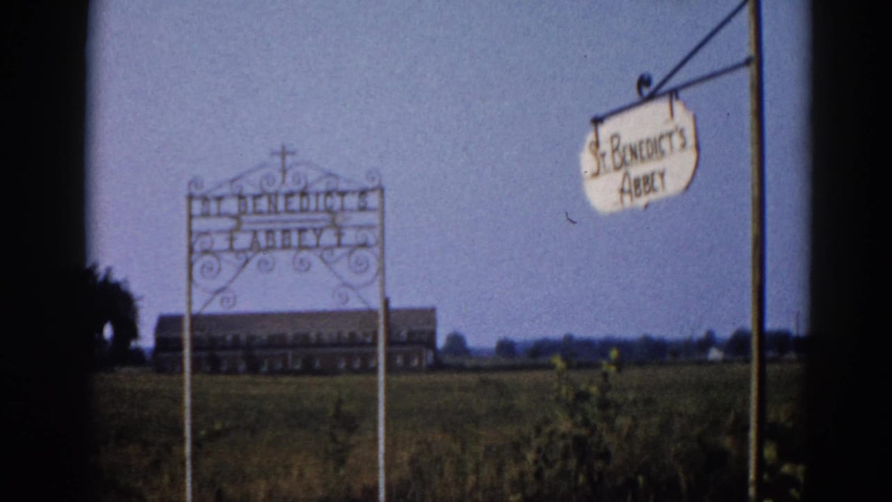 Static 8mm shot of St. Benedict's Abbey entrance signage with ornate ironwork scrollwork and distant brick institutional building; Illinois, c.1962.