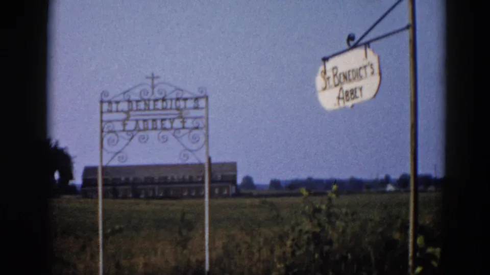 Closing frame of static 8mm shot; wrought-iron St. Benedict's Abbey sign and pendant sign against pale sky with large brick abbey building in background; Illinois, 1962.