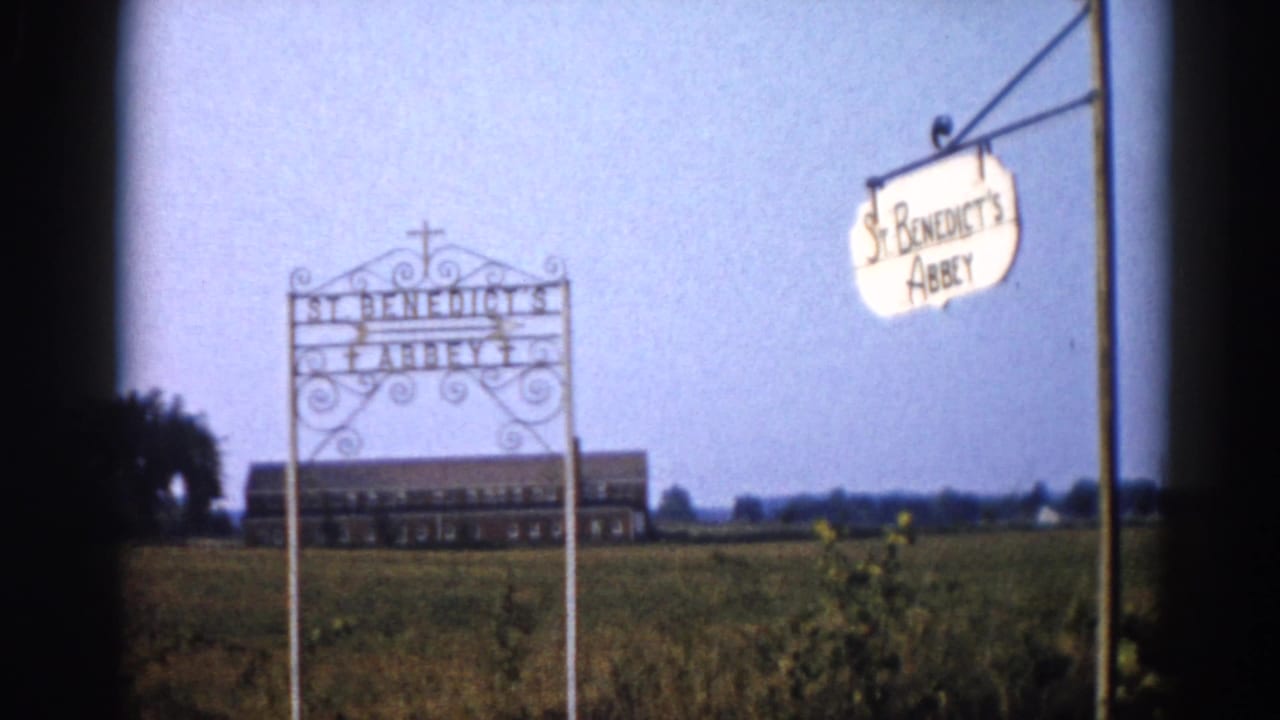 Exterior of St. Benedict's Abbey, Illinois, 1962 — wrought-iron entrance arch with cross and hanging oval sign, brick abbey building visible across flat rural fields under clear blue sky.