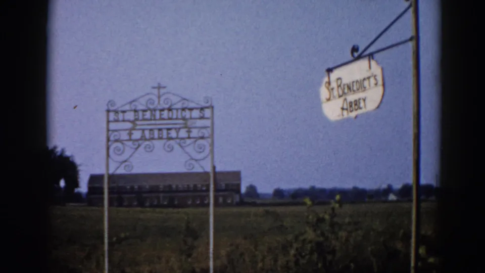 1962 8mm frame of St. Benedict's Abbey, Illinois: wrought-iron gate arch and hanging oval sign in foreground, abbey buildings on the flat rural horizon under blue sky.