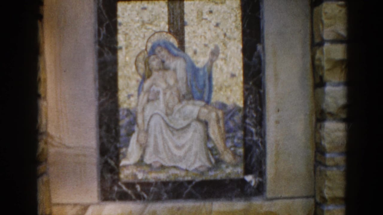 Pietà mosaic on a dark-framed panel set against a stone church wall; Virgin Mary in blue robe cradles Christ against a gold tesserae background.