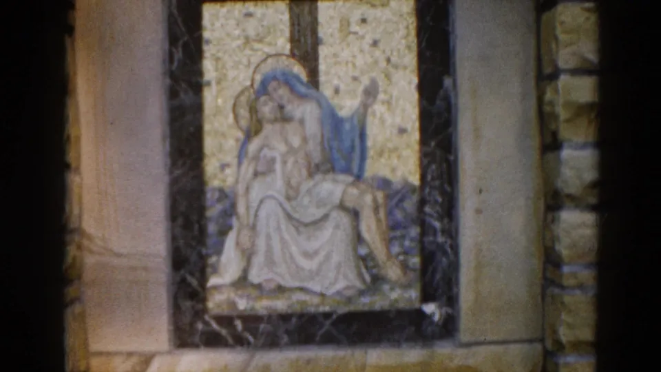 1962 8mm home-movie frame of a Pietà mosaic — Virgin Mary in blue robe cradling Christ on a gold tesserae ground — in a dark-framed panel within a stone-walled Illinois church interior.