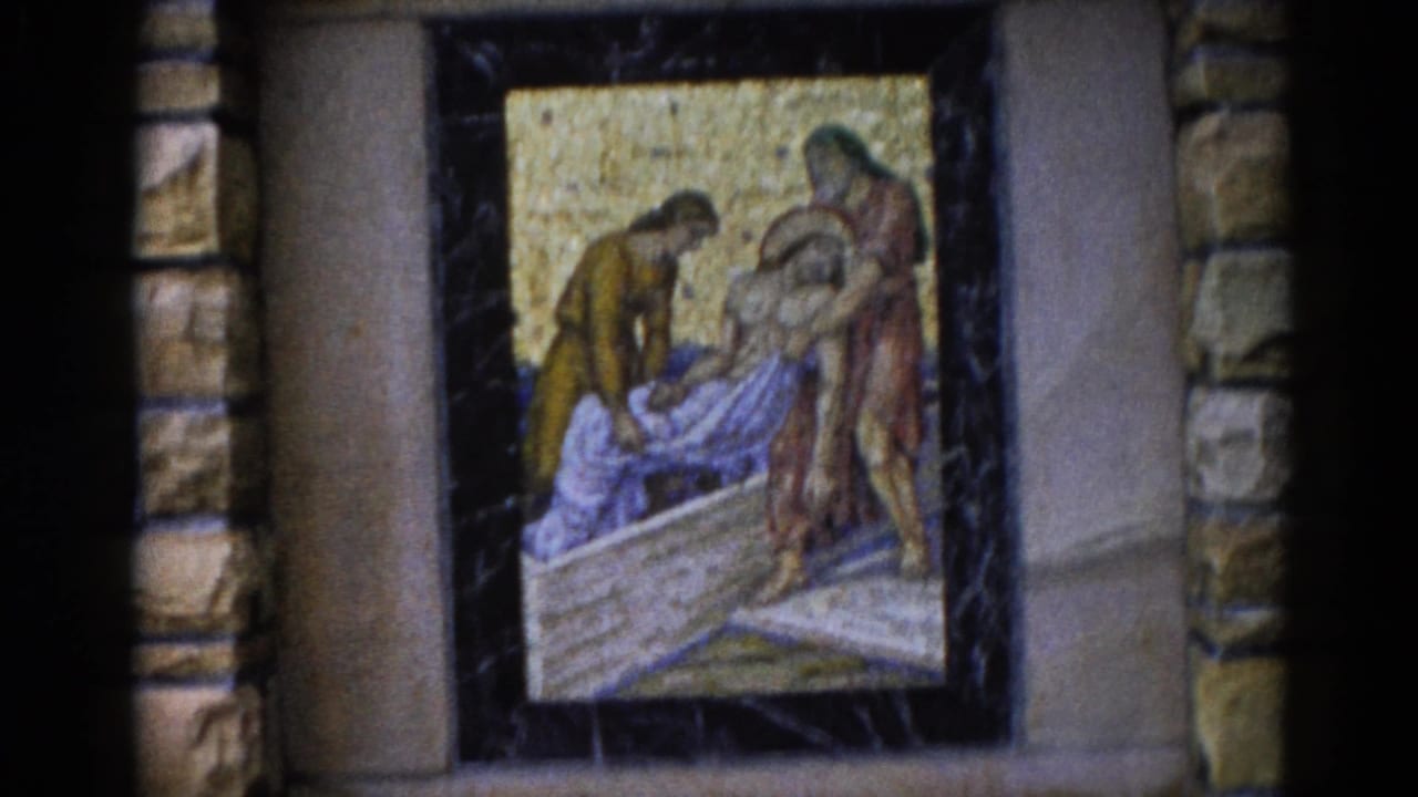 Deposition mosaic on a dark-framed panel against a stone wall; robed figures lower a body onto a stone slab with a gold tesserae background.