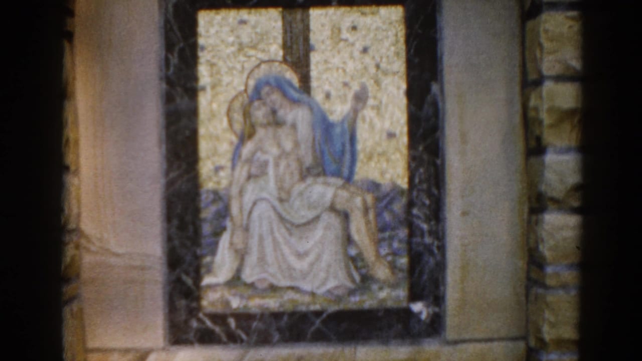 Mosaic Pietà panel showing blue-robed Mary cradling white-robed Christ, set in a dark marble-framed niche with flanking stone church walls.