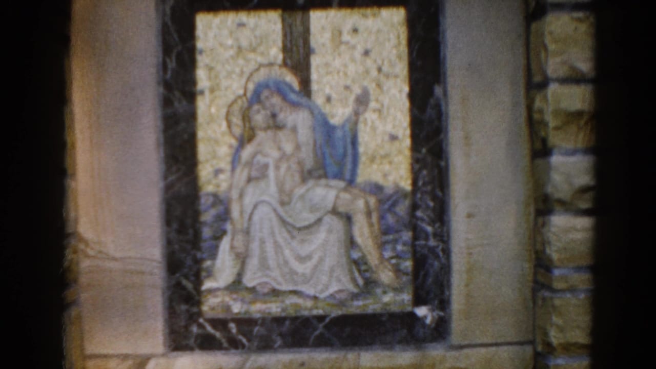 Same Pietà mosaic in dark marble frame; camera has shifted slightly, exposing more of the right stone church wall.