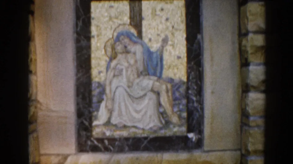 Third 8mm frame of 1962 Illinois church footage; camera has drifted leftward, exposing more of the cut-stone wall flanking the dark marble-framed Pietà.