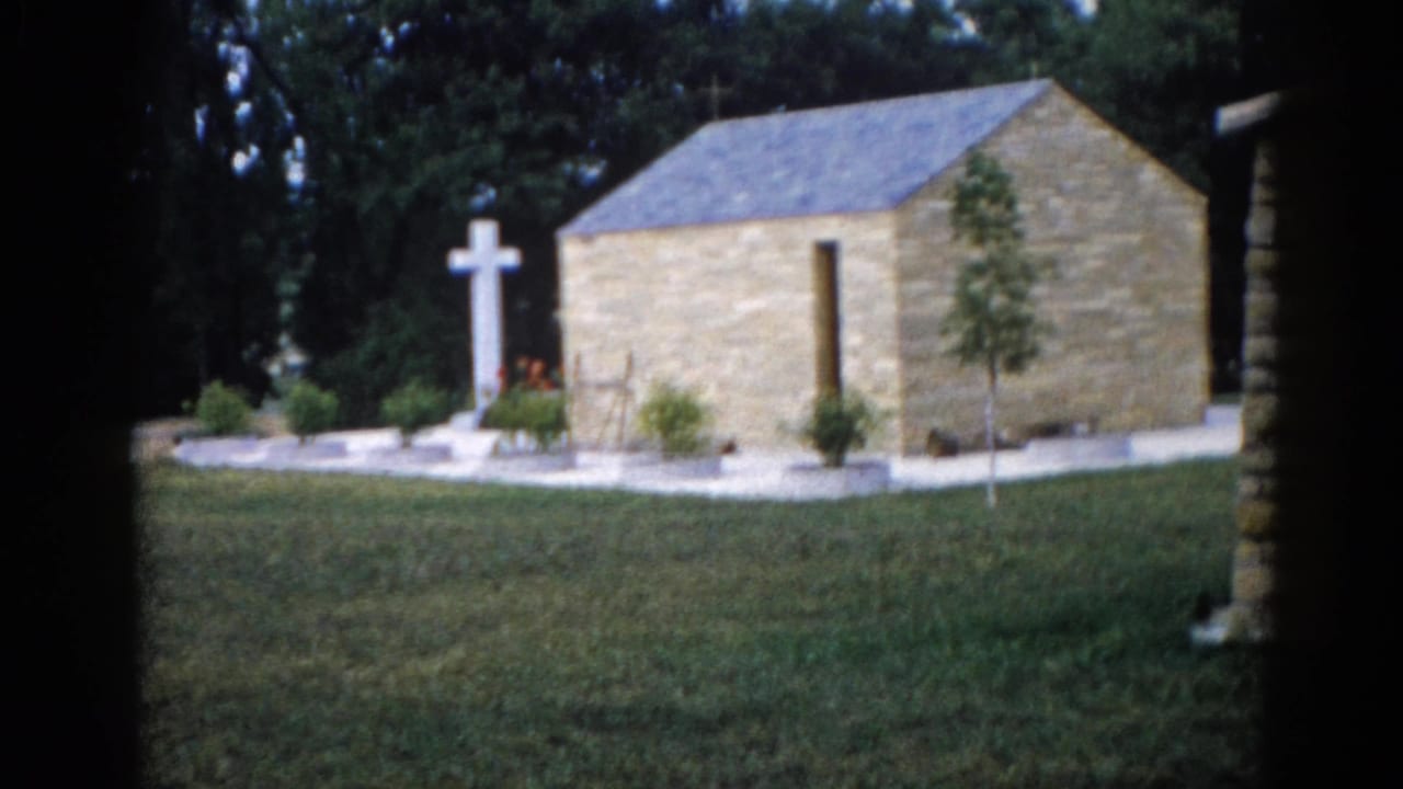 Exterior of a small stone chapel with slate roof and white freestanding cross, photographed on 8mm film in Illinois, c.1962.