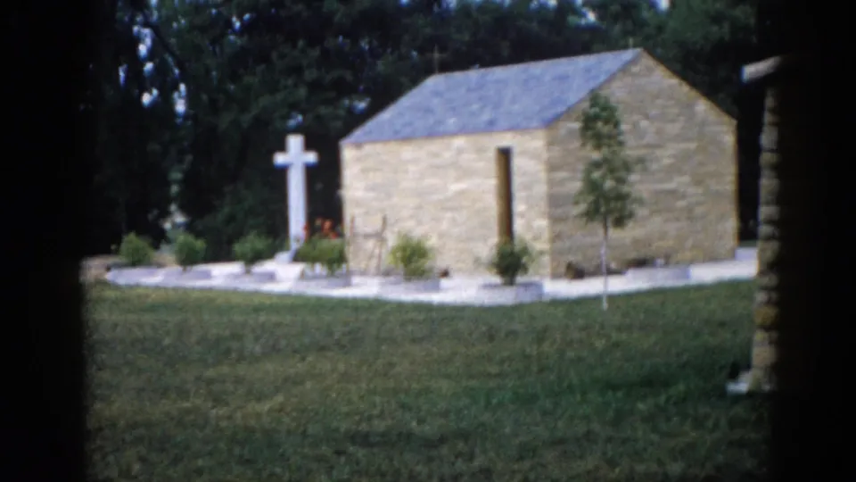 Exterior of a small stone chapel with slate roof and white freestanding cross, photographed on 8mm film in Illinois, c.1962.
