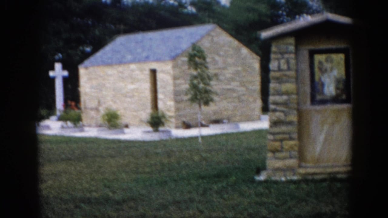 Stone chapel with gray hip roof and tall white Latin cross, religious mosaic on right-side pillar, young trees and flower beds.