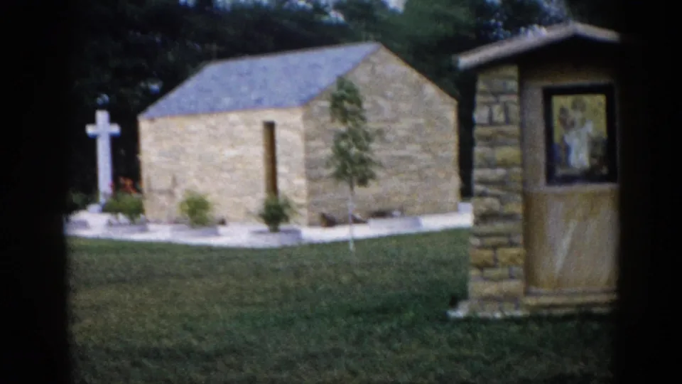 Outdoor stone chapel with white Latin cross and religious mosaic panel visible on stone pillar, planted grounds, Illinois, 1962, 8mm home movie.