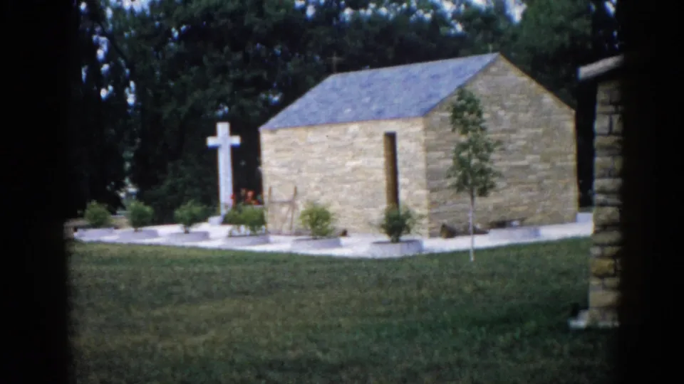 Wide establishing shot of outdoor stone chapel grounds with white Latin cross, young saplings, and planted flower beds, Illinois, 1962, 8mm home movie.