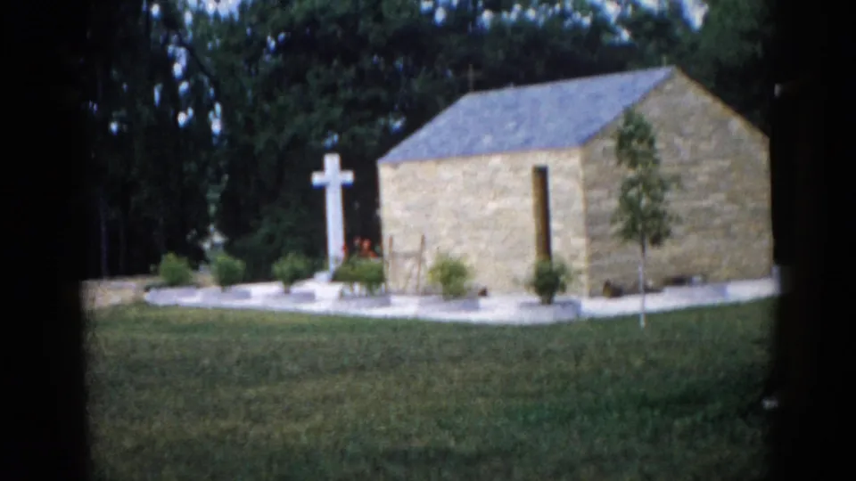 Stone chapel exterior with white Latin cross, stone pillar, and landscaped grounds including flower beds, Illinois, 1962, 8mm home movie.