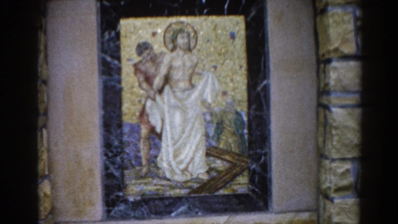 Mosaic panel in dark marble frame on cut-stone wall, depicting a halo-bearing robed figure alongside a second figure and a diagonal cross element.