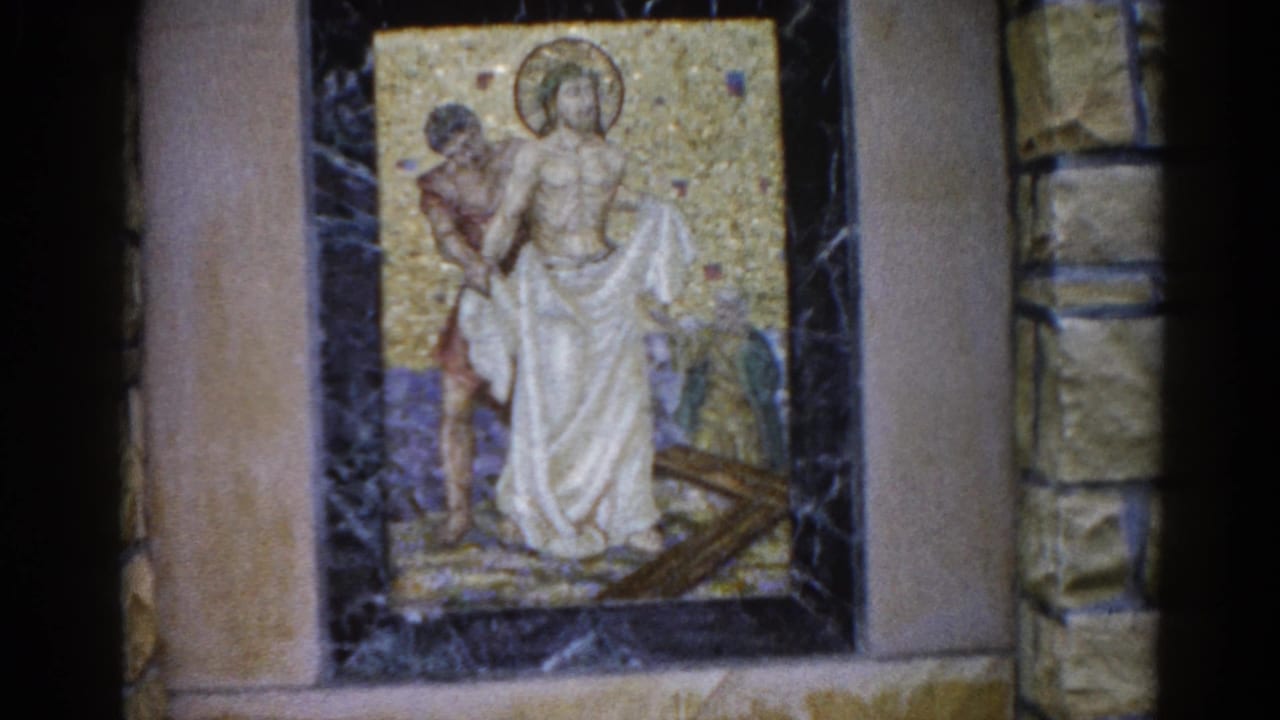 Mosaic on stone wall with halo-bearing robed figure and companion beside a cross; camera has shifted slightly, exposing more of the right stone column.