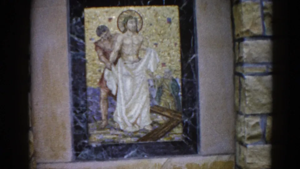 8mm film frame, 1962, Illinois: mosaic panel with halo figure and cross element; stone wall more visible at top of frame as camera settles on a lower angle.