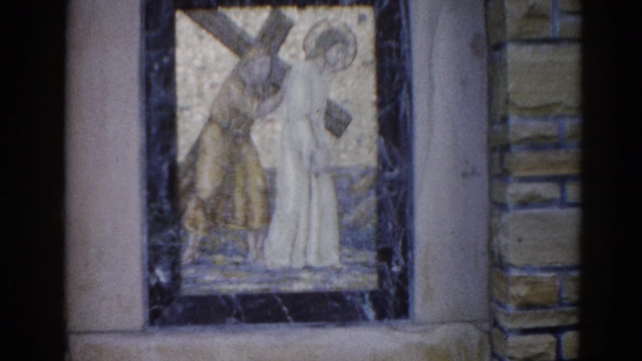 Mosaic panel showing two robed figures — one haloed carrying a cross — framed in dark marble against a stone church wall.