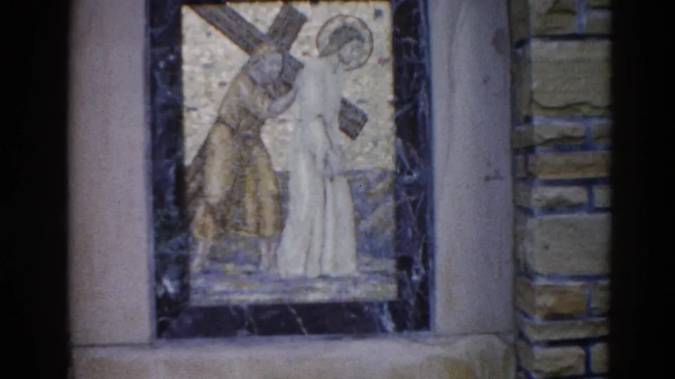 Closer 8mm frame from a 1962 Illinois church mosaic depicting Jesus (halo, white robe) beneath a cross with a companion figure, gold tessera ground.
